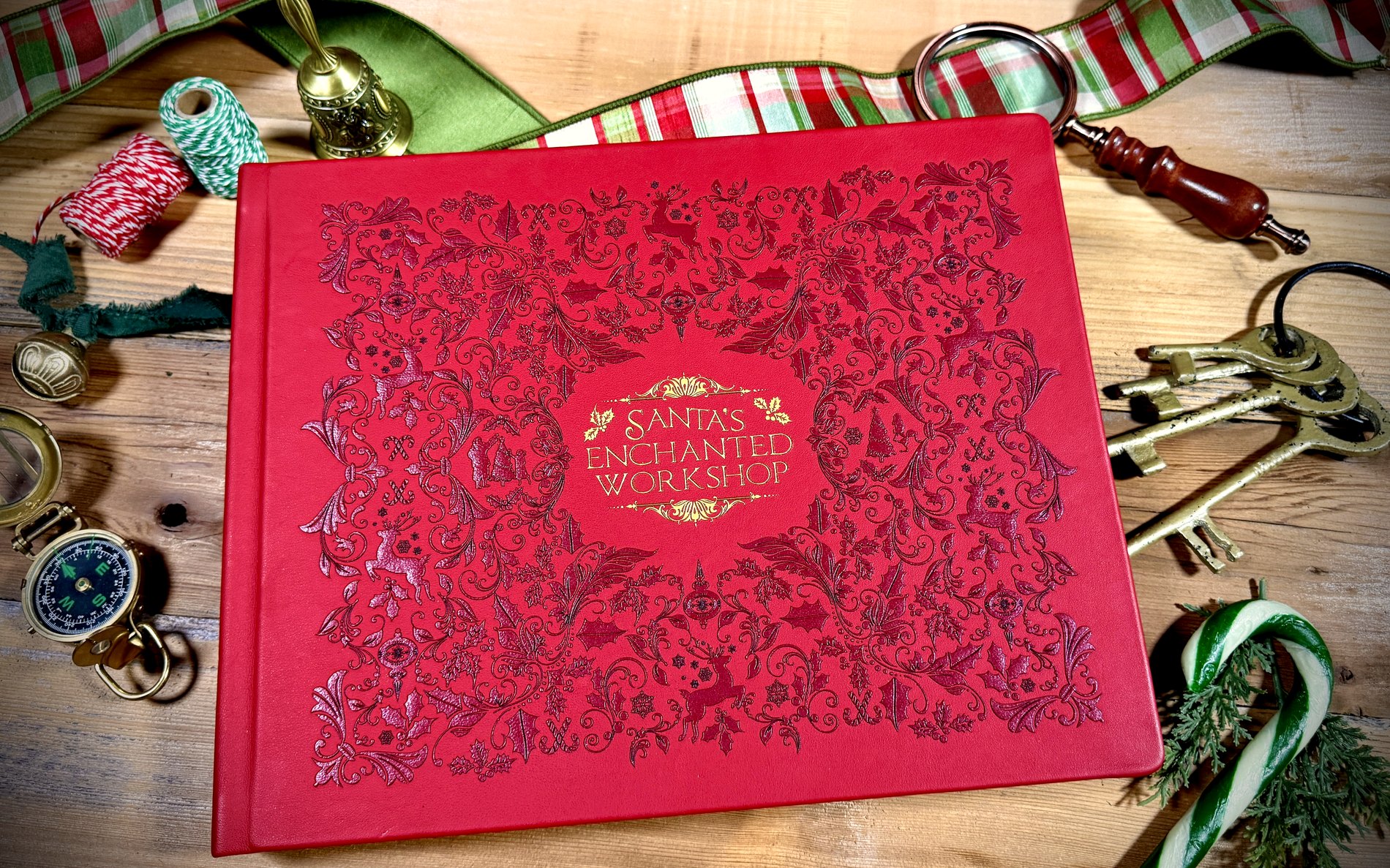 The red leather keepsake album embossed “Santa’s Enchanted Workshop”, on the workshop table with brass keys, a compass and a magnifying glass